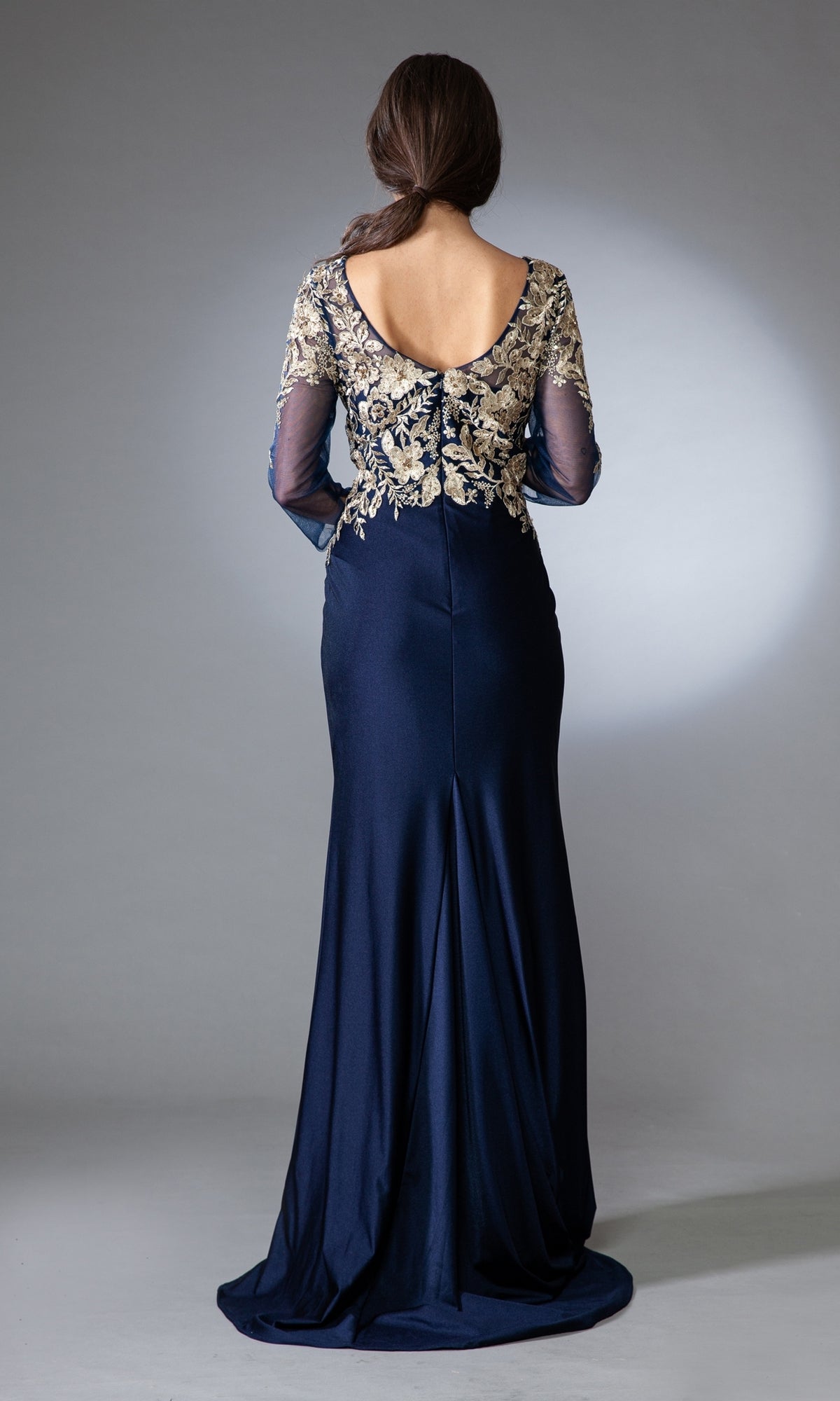 A woman stands facing away in an Amelia Couture 7039 navy mother-of-the-bride dress, featuring sheer long sleeves and gold floral embroidery on the upper back and arms, set against a plain gray background.
